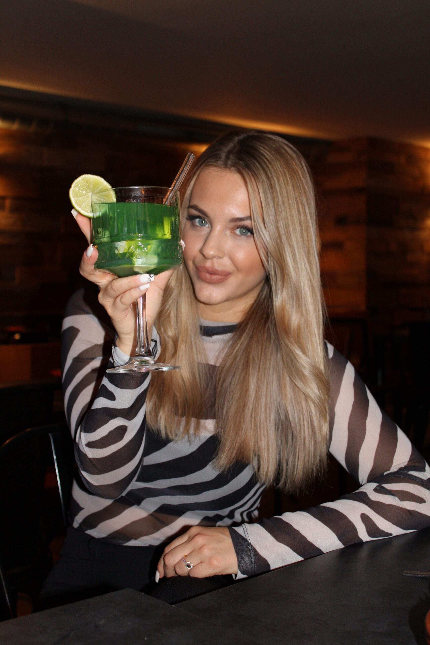 Frau mit langem blondem Haar hebt einen grünen Cocktail mit Limette im Tapas & More Dortmund.