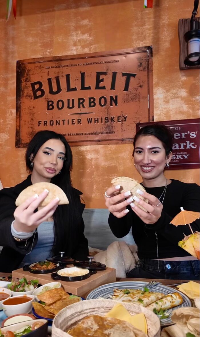 Zwei Frauen halten Tacos in die Kamera im Tequilaria Bochum vor rustikaler Wand mit Bulleit-Bourbon-Schild
