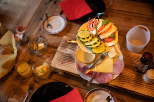 Frühstücksetagere mit Käse, Wurst, Gurke, Tomate und Obst auf rustikalem Holztisch
