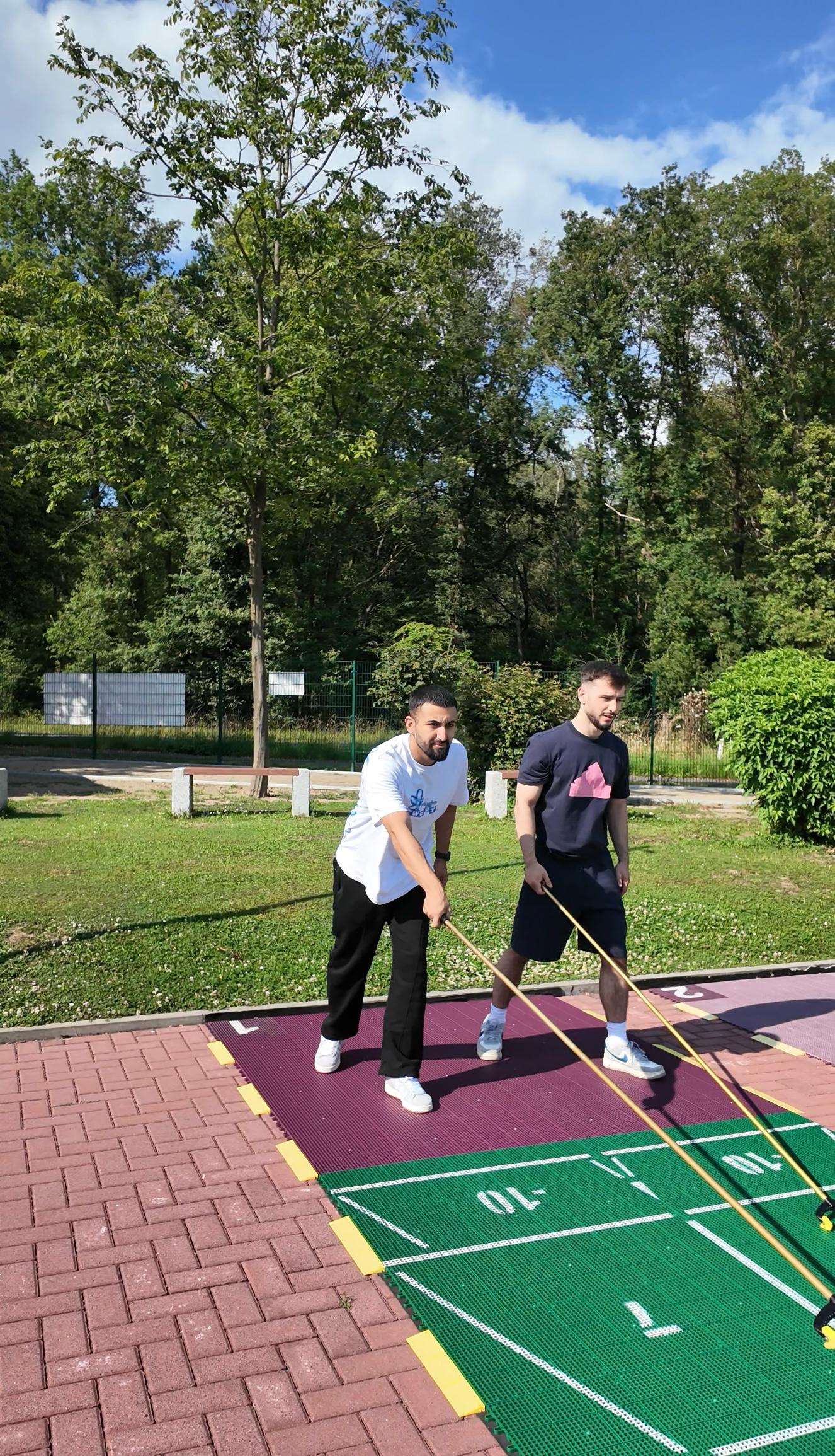 Zwei junge Männer machen gemeinsam ein Teamspiel auf Holzskiern im Sport- und Freizeitpark Limburg-Linter, umgeben von Grünanlagen und guter Laune