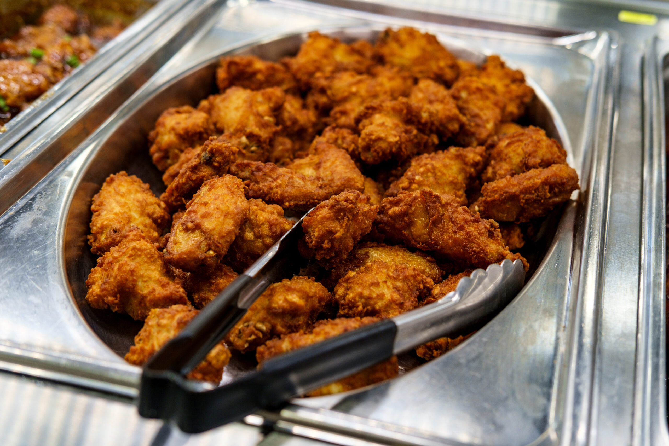 Nahaufnahme von knusprigen, goldbraun gebackenen Chicken Wings, die im Rahmen des amerikanischen All-You-Can-Eat-Buffets von ISM Foodspot Dortmund serviert werden. Eine Servierzange liegt bereit, um die Wings bequem aufzunehmen.
