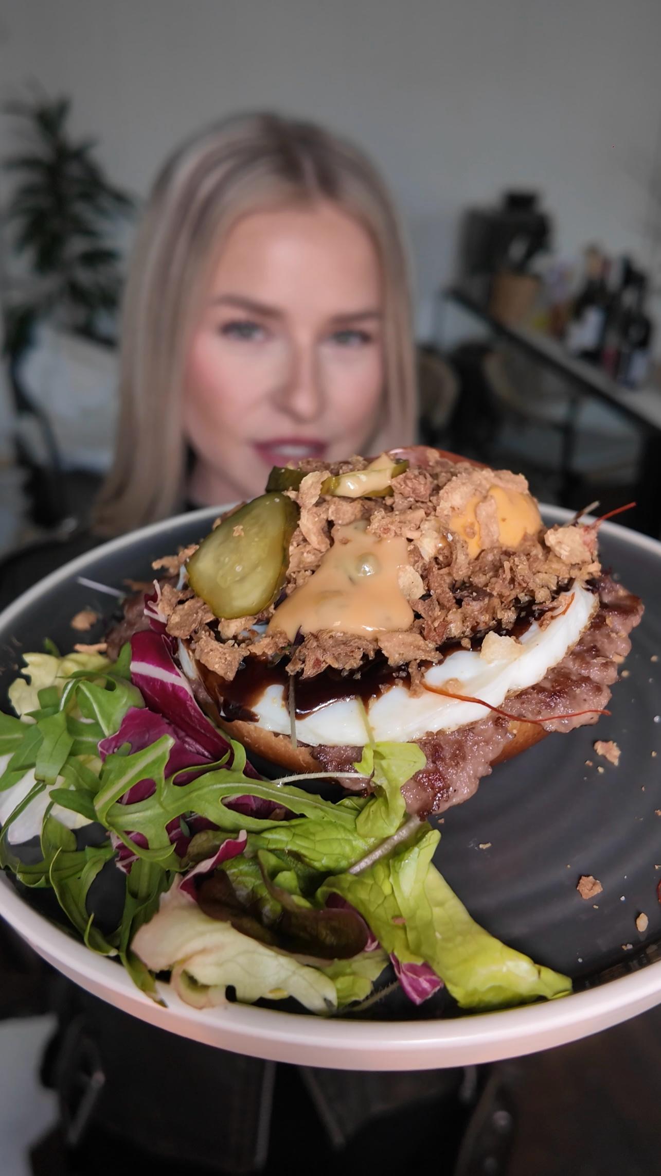 Nahaufnahme eines LALA Smashburgers in Frankfurt – serviert mit saftigem Patty, eingelegten Gurken, Zwiebeln und frischem Salat, im Hintergrund eine Frau
