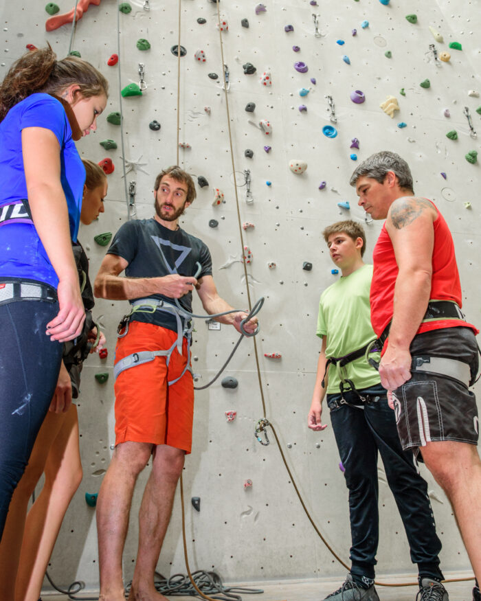 Trainer erklärt einer Gruppe von Kletternden den Umgang mit Seil und Sicherung in der MOVE Kletterhalle vor einer hohen Kletterwand