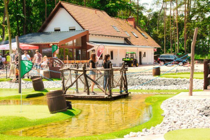 Übersicht über den Adventure Golf Bereich im Sport- und Freizeitpark Limburg-Linter mit Holzbrücke, Wasserlauf und Hauptgebäude im Hintergrund