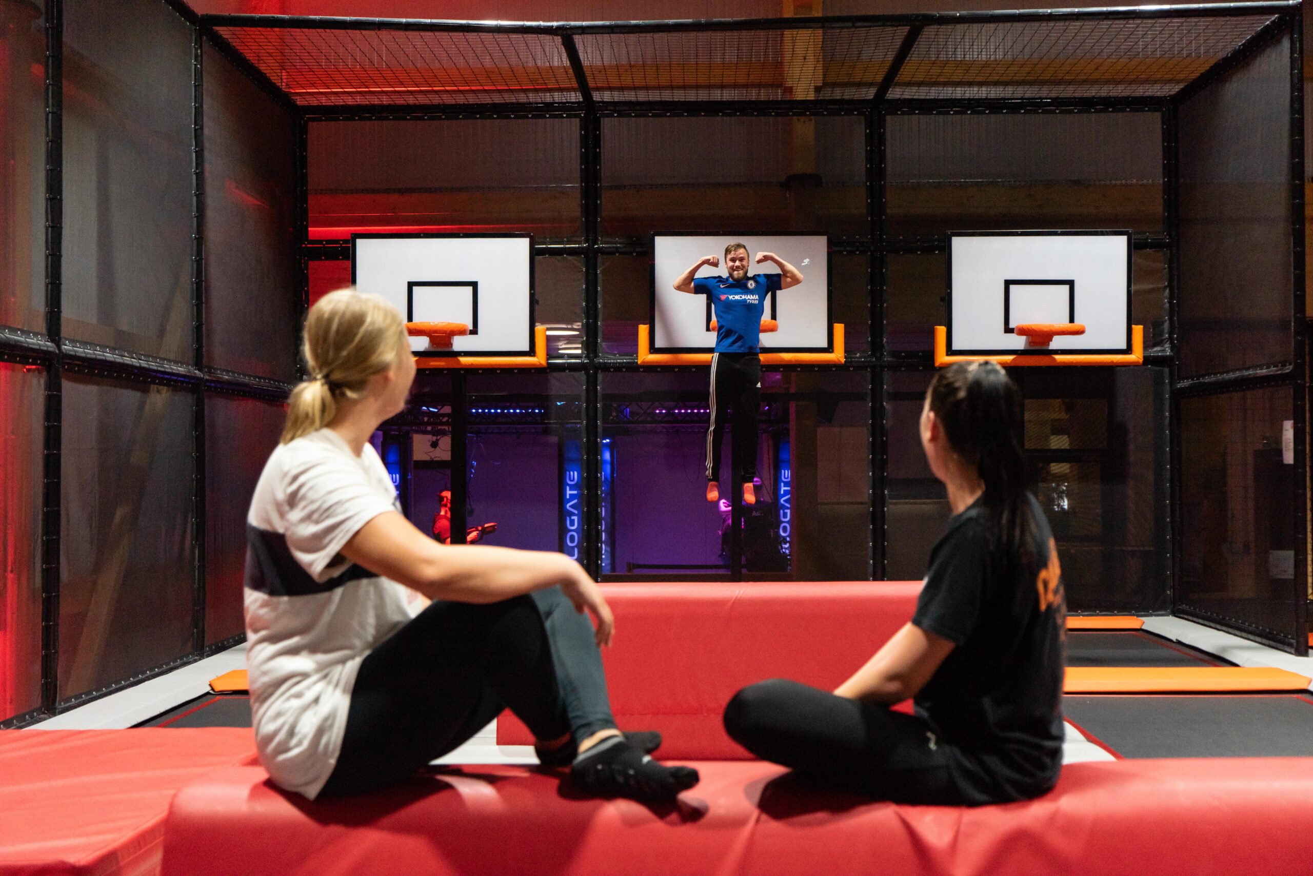 Person springt auf einem Trampolin und trifft einen Basketballkorb in der Freizeitwelt Sauerland, während zwei Zuschauer auf einem roten Kissen sitzen.