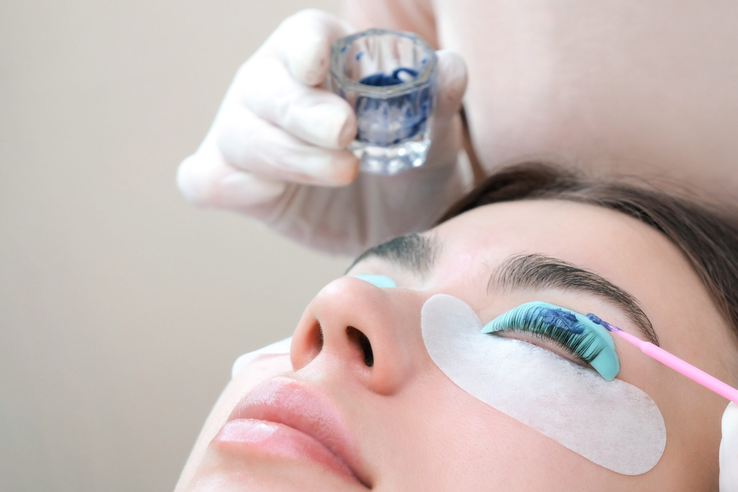 Nahaufnahme des Gesichts einer Frau während einer Wimpernfärbung. Eine Kosmetikerin hält ein kleines Glas mit blauer Wimpernfarbe bereit, während die Augenlider der Frau mit Schutzpads abgedeckt sind.