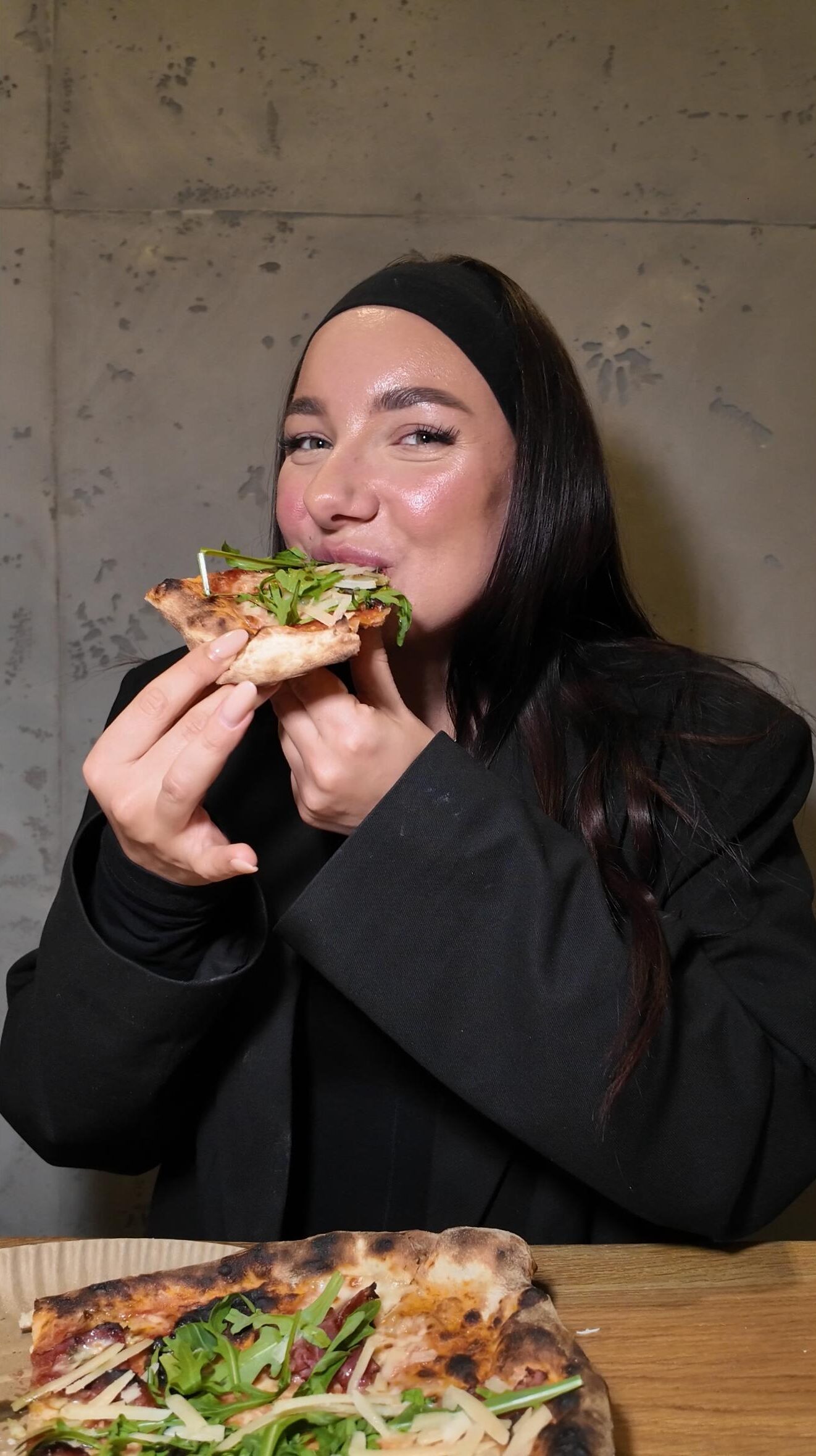 Eine Frau genießt lächelnd ein Stück Pizza, belegt mit frischem Rucola und Tomaten, bei Zampano To Go in Frankfurt