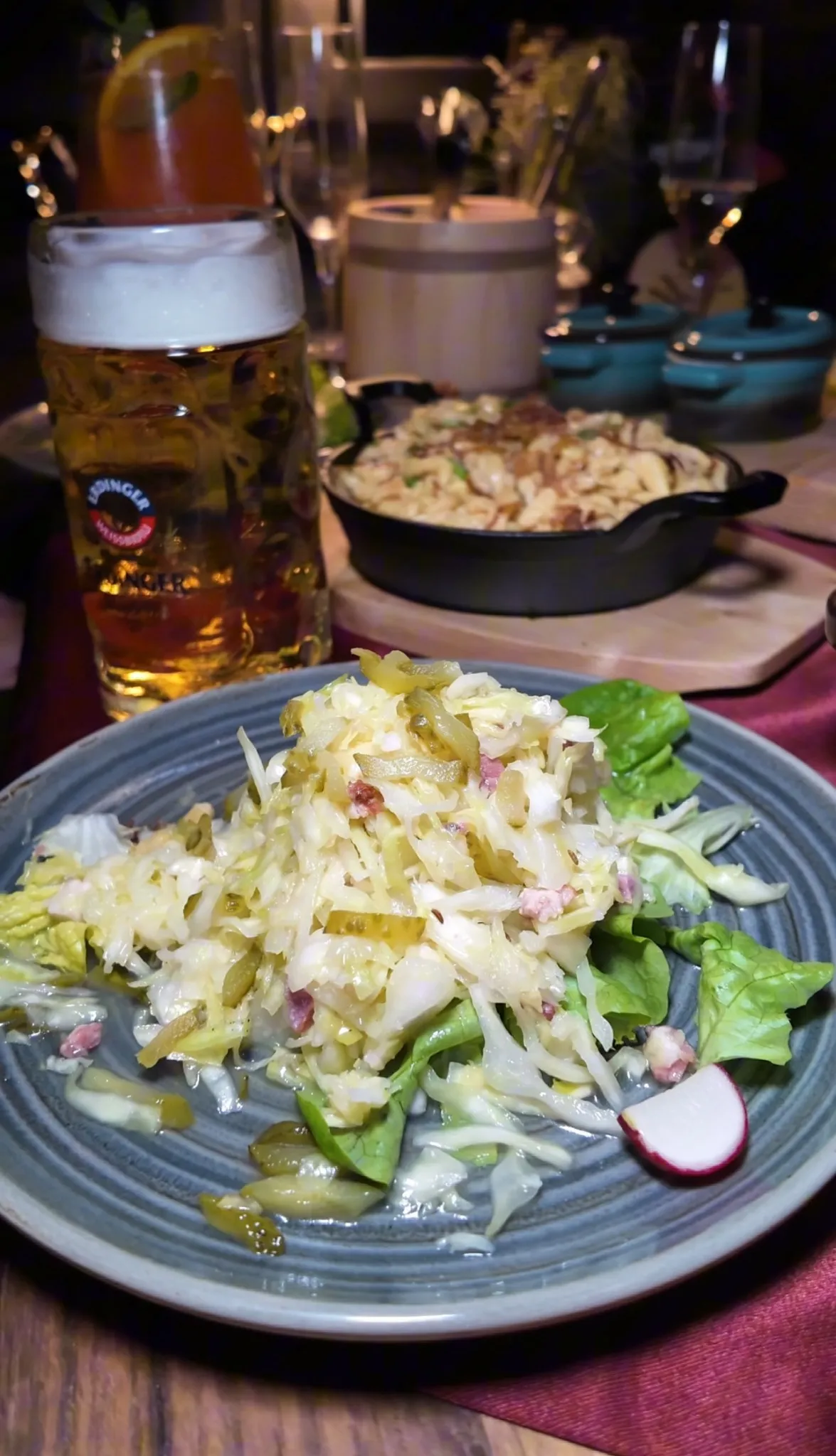 Typischer Alm-Teller in der Alm Fernweh mit Krautsalat auf Blattsalat, dahinter Käsespätzle in der gusseisernen Pfanne und ein Maß Bier auf rustikal gedecktem Tisch