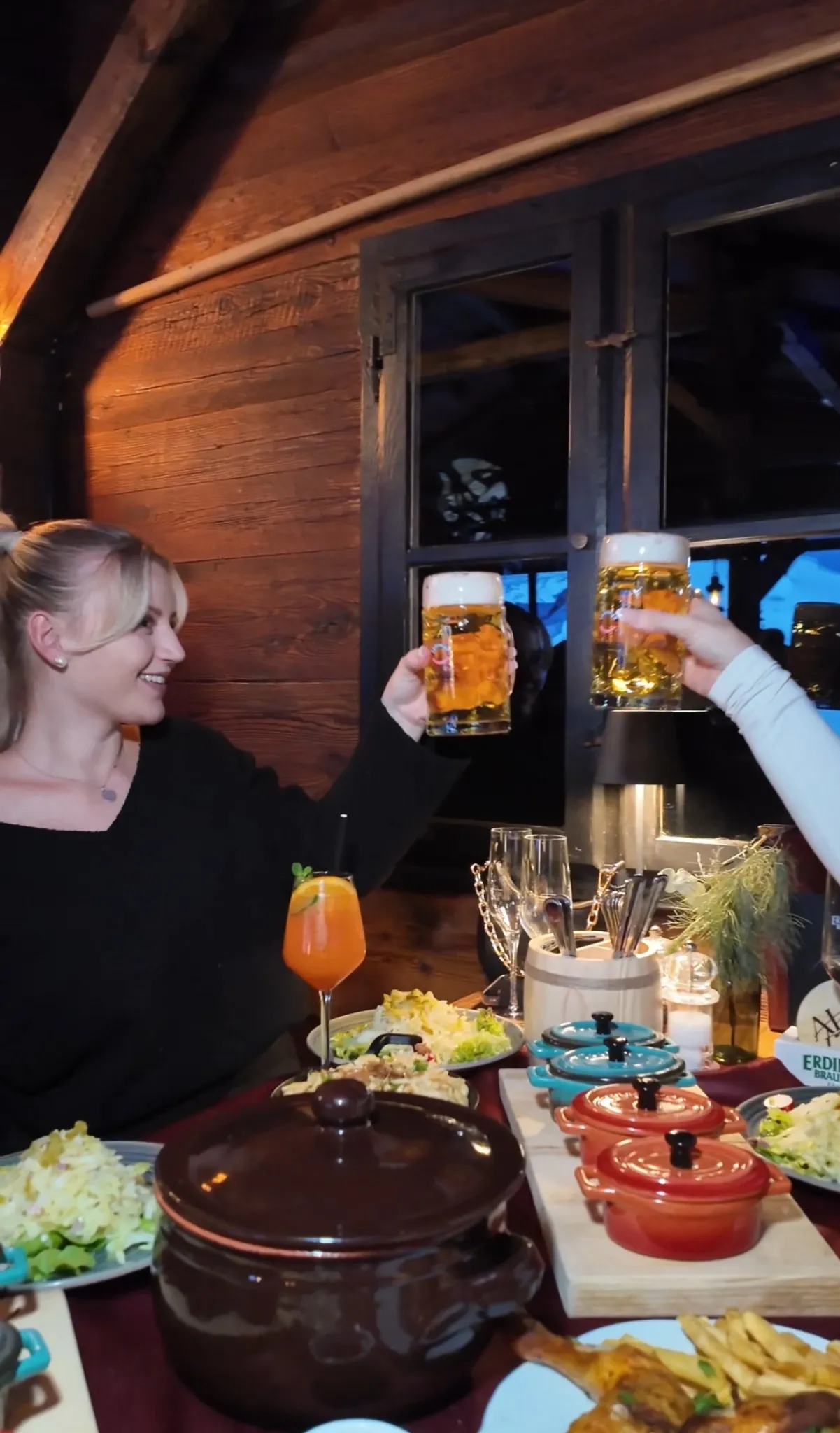 Zwei Personen stoßen mit Bierkrügen an, auf dem Tisch stehen Käsespezialitäten, Cocktails, Kerzen und typische Hüttengerichte in der Alm Fernweh