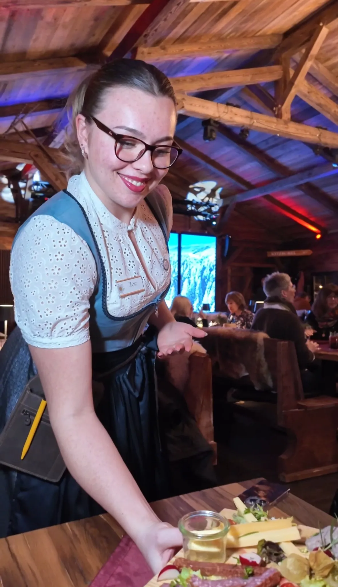 Lächelnde Servicemitarbeiterin in Dirndl serviert Speisen bei Abendveranstaltung in der Alm Fernweh, im Hintergrund rustikales Ambiente mit Holzbalken und Gästen