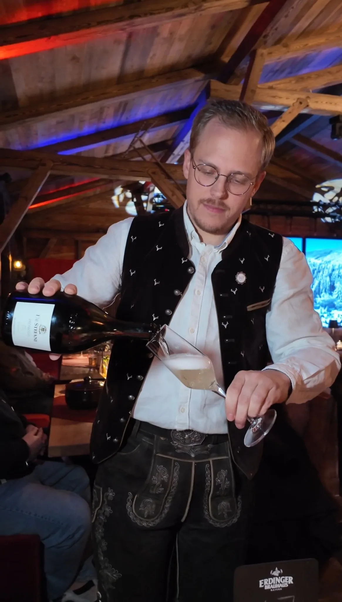 Servicemitarbeiter in traditioneller Tracht schenkt stilvoll Wein ein bei Abendveranstaltung in der Alm Fernweh, im Hintergrund rustikale Almhütten-Atmosphäre