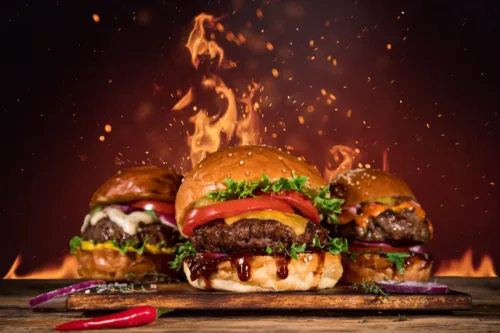 Drei saftige Burger mit verschiedenen Toppings wie Käse, BBQ-Sauce, Salat und Zwiebeln, im Hintergrund lodern Flammen für einen feurigen Effekt