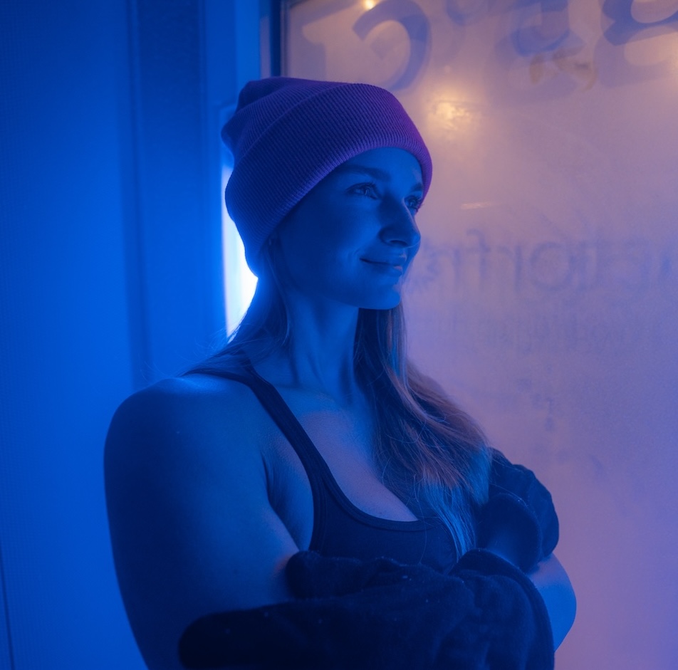 Frau mit Mütze und Sportkleidung steht in der vereisten Kryokammer bei Doctorfrost Frankfurt, umgeben von blauem Licht und kondensierter Kälte an der Glasscheibe