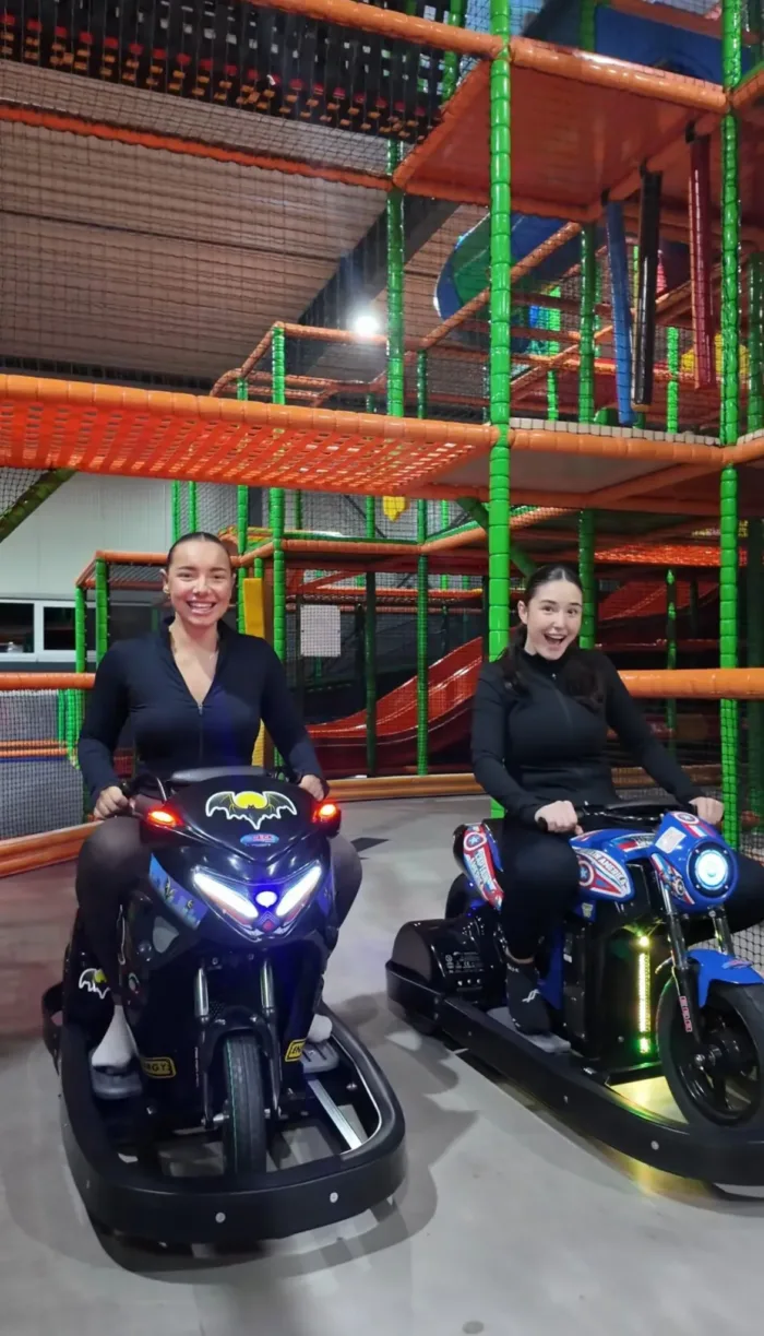 Zwei Personen sitzen lachend auf elektrisch betriebenen Kinder-Motorrädern im FunFusion Wetzlar, im Hintergrund ein bunter Indoor-Kletterbereich