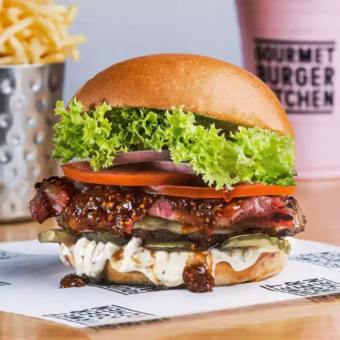 Gourmet-Burger mit Bacon, Salat, Tomate, Zwiebel, Gurke, cremiger Sauce und fluffigem Bun, im Hintergrund Pommes und Milchshake