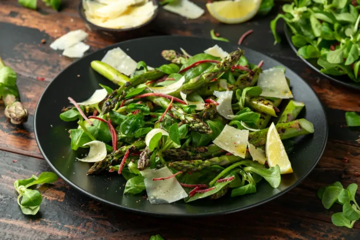 Frischer grüner Spargelsalat mit Rucola, Parmesanraspeln und Zitronenspalten auf einem dunklen Teller angerichtet