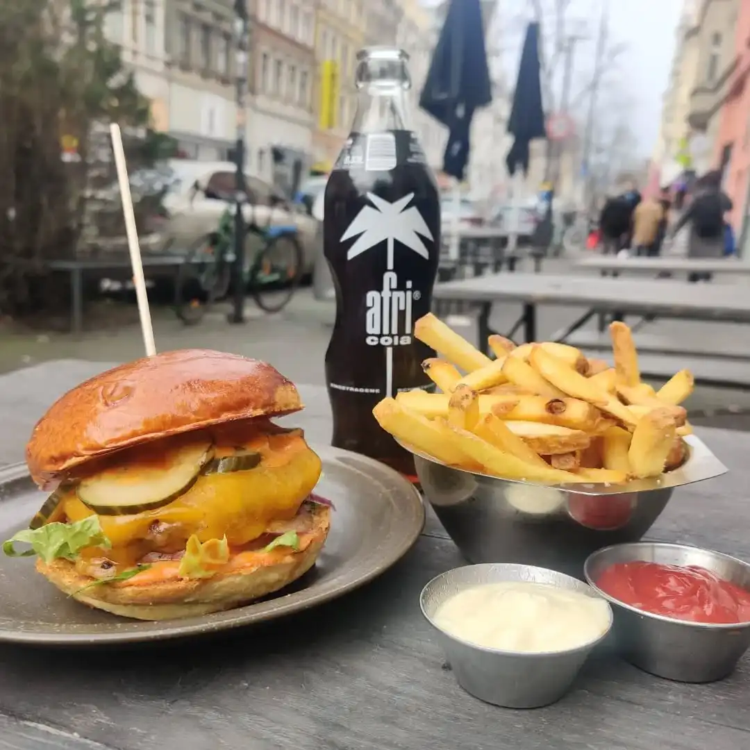 Cheeseburger mit Pommes und Afri Cola auf einem Holztisch vor dem Hornochse Köln, serviert mit Dips und Blick auf die Außengastronomie der Straße