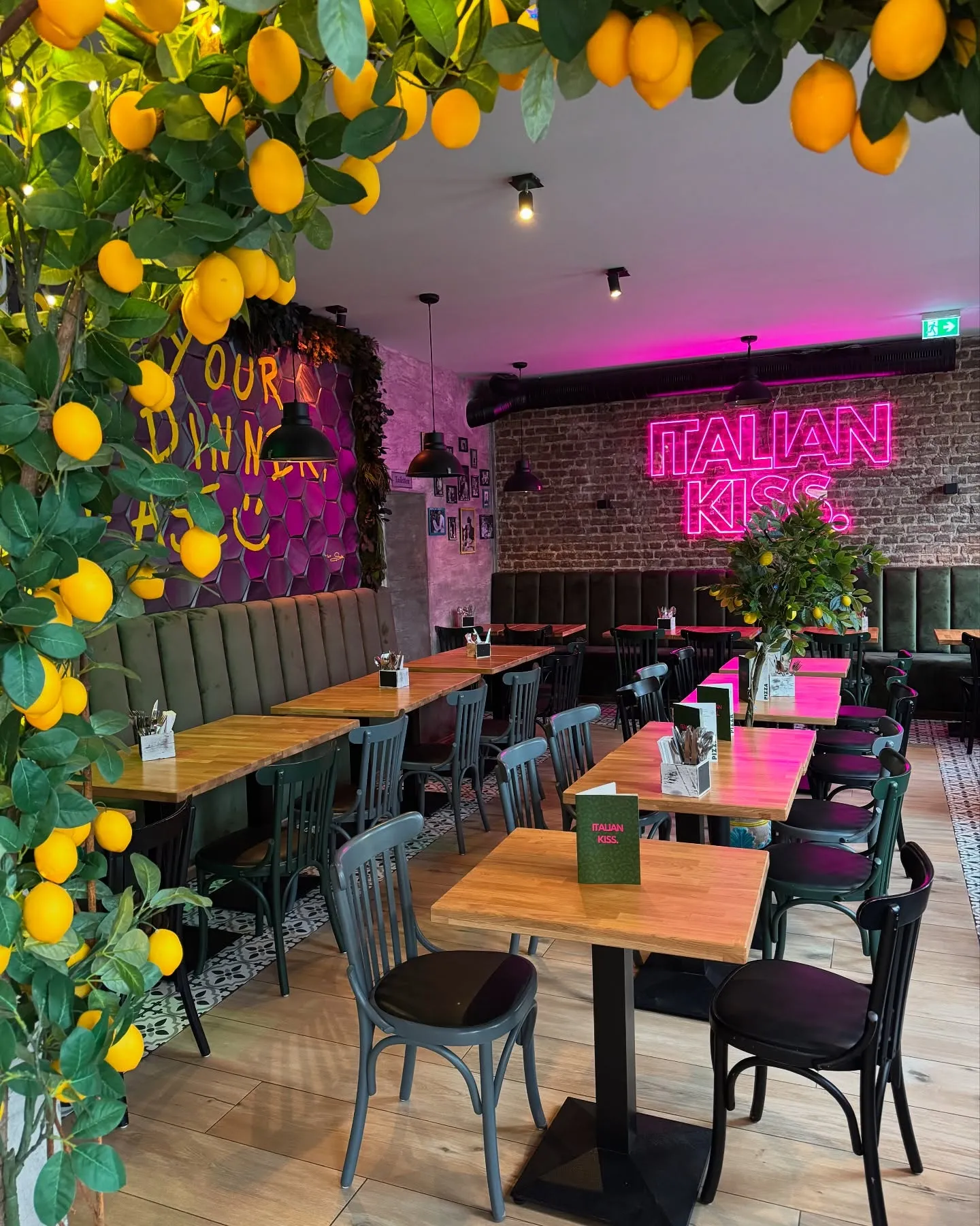 nnenraum des Italian Kiss Köln mit langen Holztischen, Zitronen-Dekoration, Pflanzen und pinker Neonschrift „Italian Kiss“ an der Wand