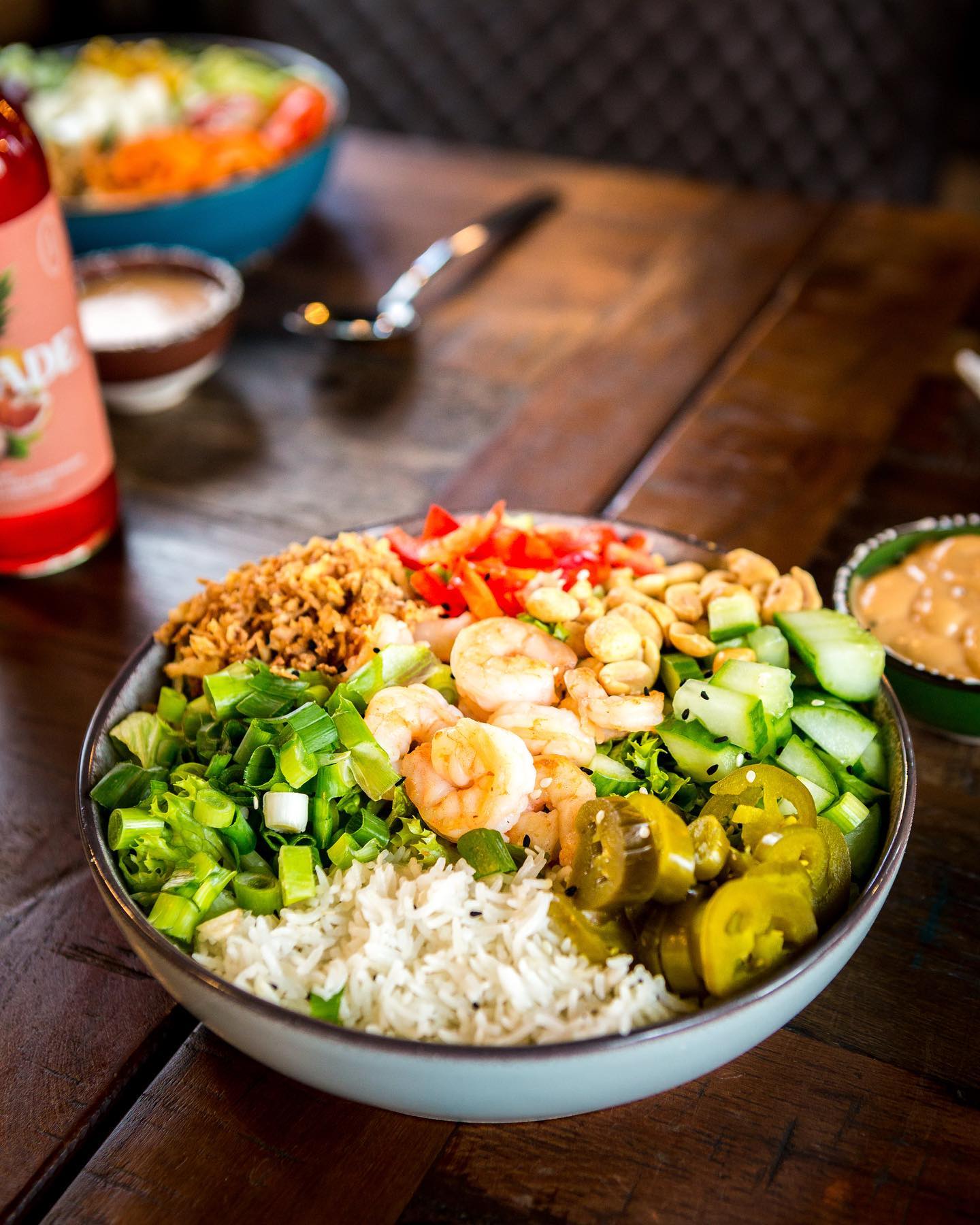 Frische Poké Bowl im Monument Burger Leverkusen mit Garnelen, Reis, Salat, Gurke, Tomate, Edamame, Pickles und Toppings, serviert mit Dip und Fritz-Schorle im Hintergrund