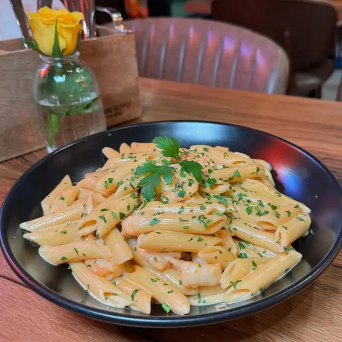 Penne in cremiger Sahnesauce, serviert in einer schwarzen Schale, garniert mit frisch gehackter Petersilie auf einem Holztisch mit gelber Rose im Hintergrund