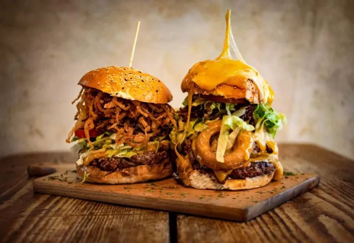 Zwei saftige BBQ-Burger auf einem Holzbrett aus dem Red Barn BBQ Restaurant, gefüllt mit Fleisch, Salat, Käse, Zwiebelringen und BBQ-Sauce