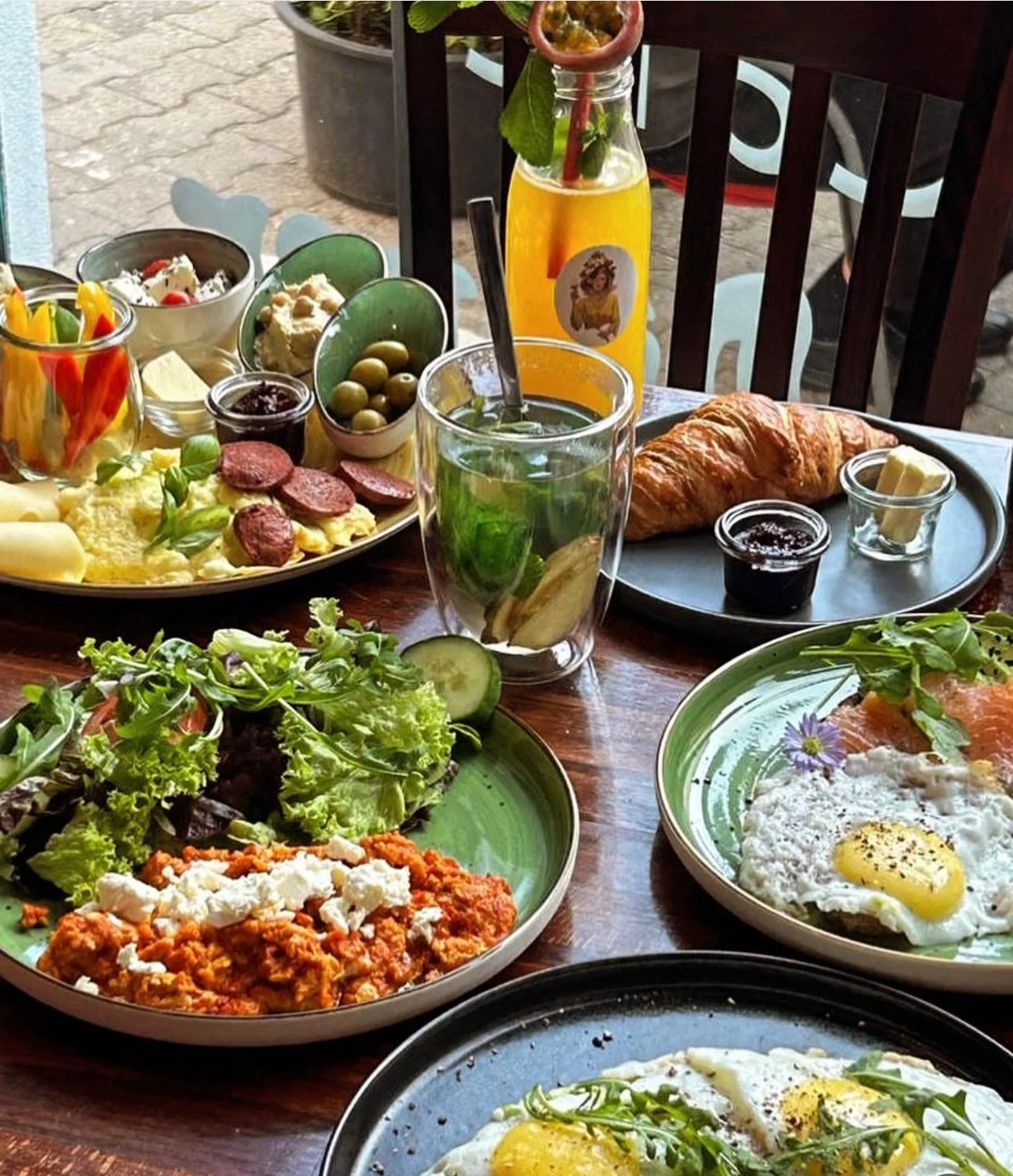Vielfältiger Brunch in der Shima Café Bar Frankfurt mit Spiegelei, Simit, Rührei, Croissant, Salat, Oliven, Aufschnitt, Obst, Minztee und Orangensaft auf rustikalem Holztisch