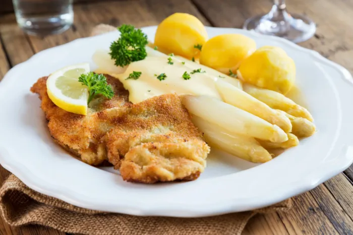 Panierte Schnitzel mit weißem Spargel, Sauce Hollandaise und Salzkartoffeln, garniert mit Petersilie und Zitrone auf einem weißen Teller