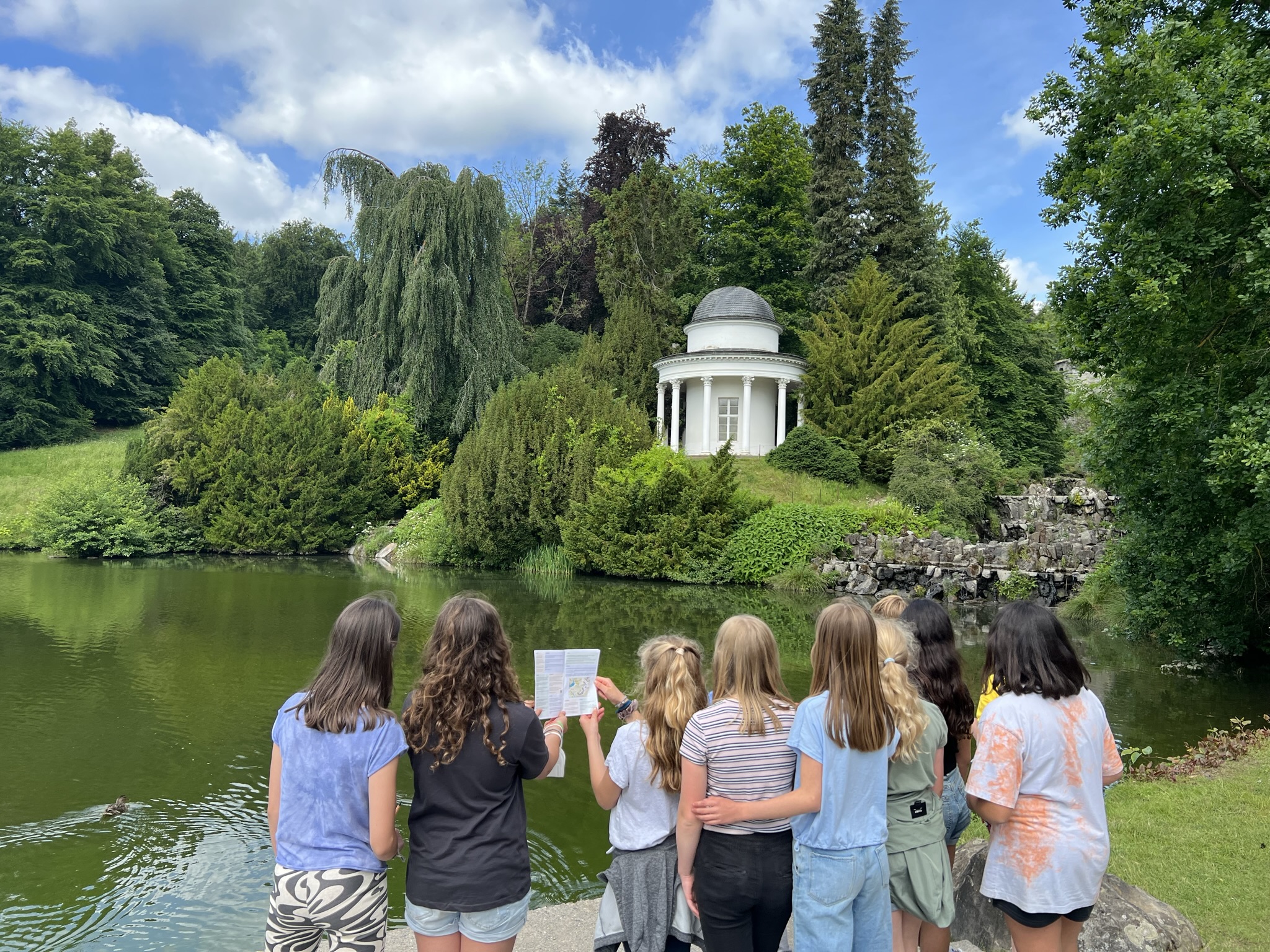 Jugendgruppe der Stadtkrimis Kassel steht mit Rätselheft am Seeufer im Bergpark und blickt auf den weißen Tempel-Pavillon, eingebettet in grüne Landschaft