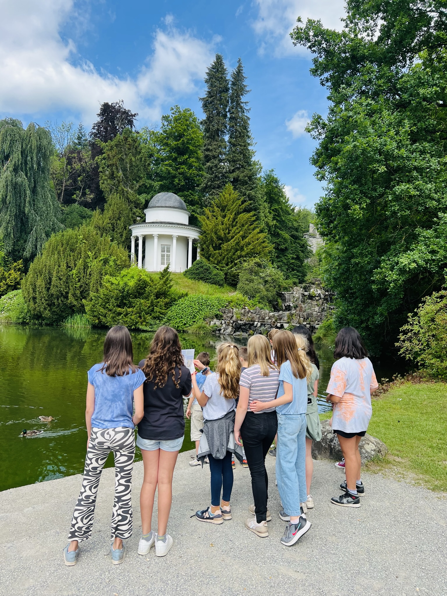 Junge Gruppe bei den Stadtkrimis Kassel steht an einem Seeufer im Park und blickt auf einen weißen Tempelpavillon, umgeben von dichter Natur im Sommer