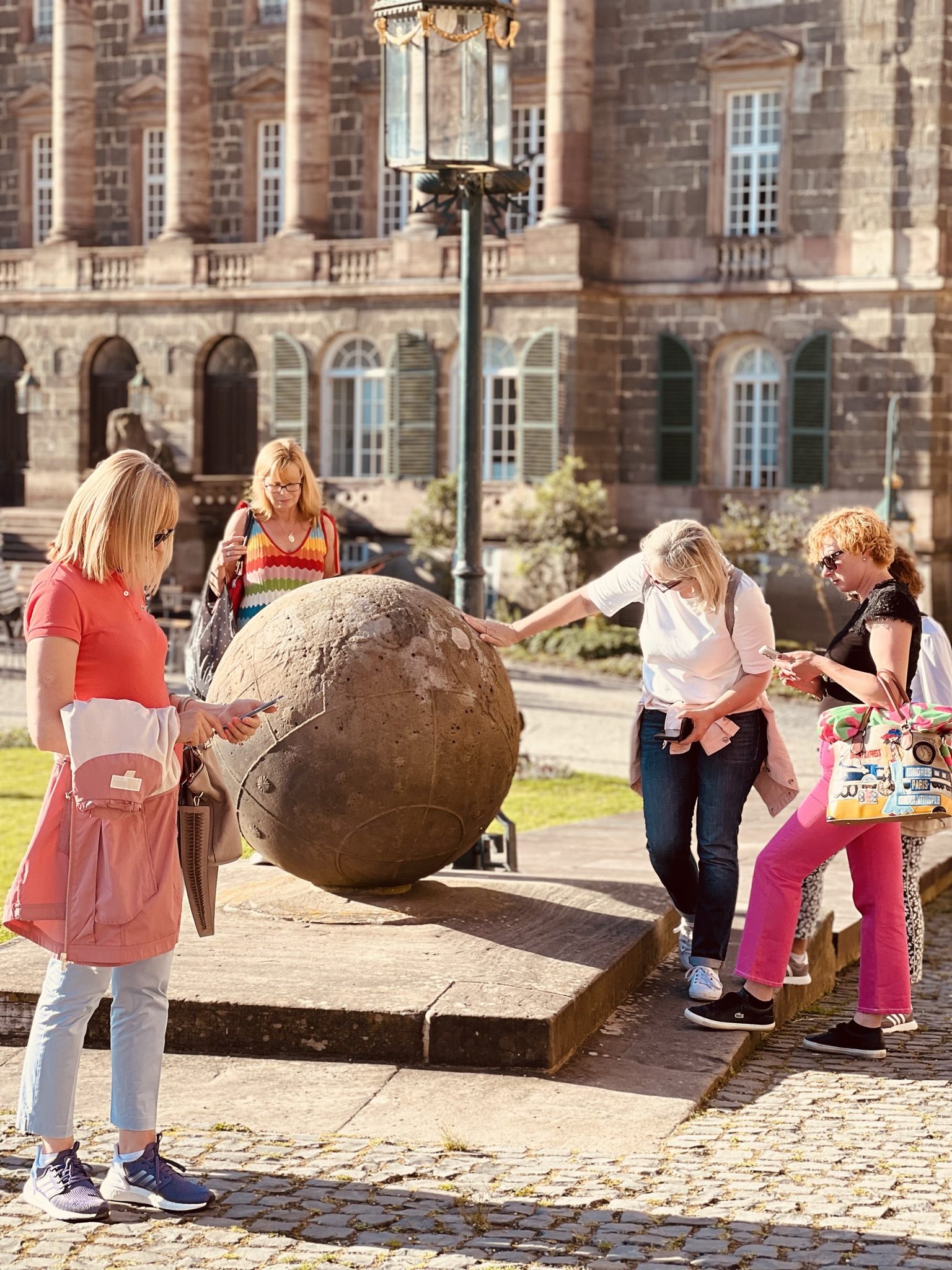Teilnehmergruppe der Stadtkrimis Kassel untersucht ein steinernes Kugel-Denkmal vor historischer Gebäudefassade bei Sonnenschein, mit Roadbook in der Hand