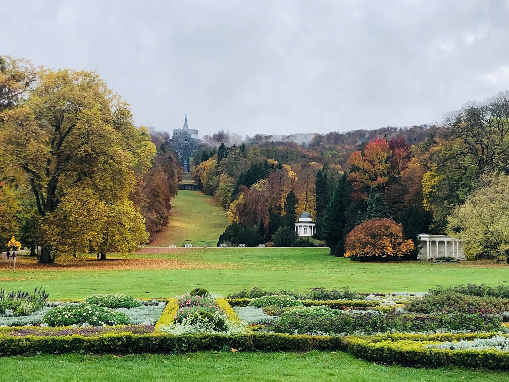 Weitläufiger Blick über den Bergpark Wilhelmshöhe im Herbst mit symmetrisch angelegtem Garten im Vordergrund, buntem Laub und der Herkulesstatue in der Ferne