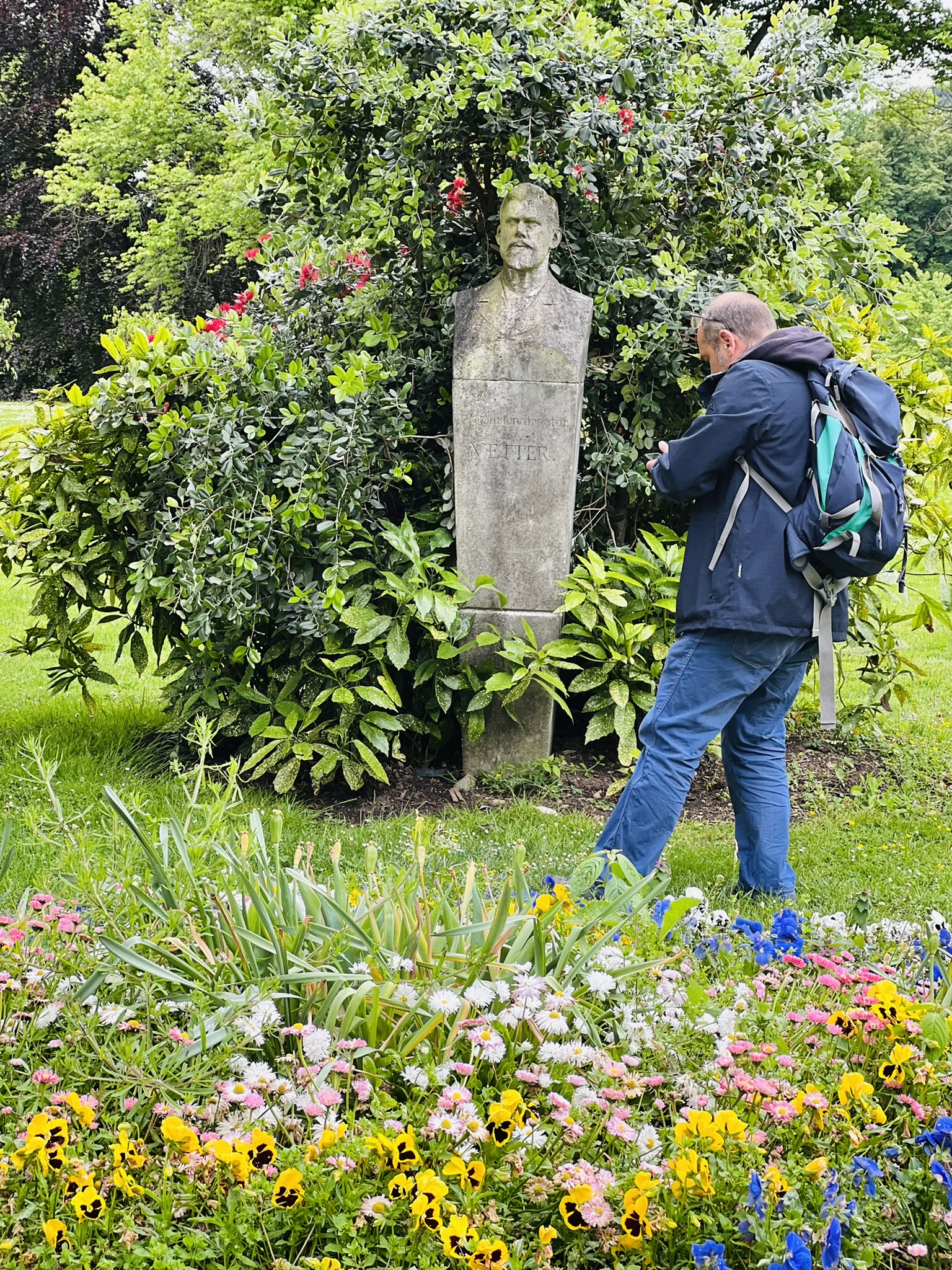 Teilnehmer der Stadtkrimis Kassel untersucht eine historische Büste im Park auf Hinweise, umgeben von Blumenbeeten und dichtem Grün