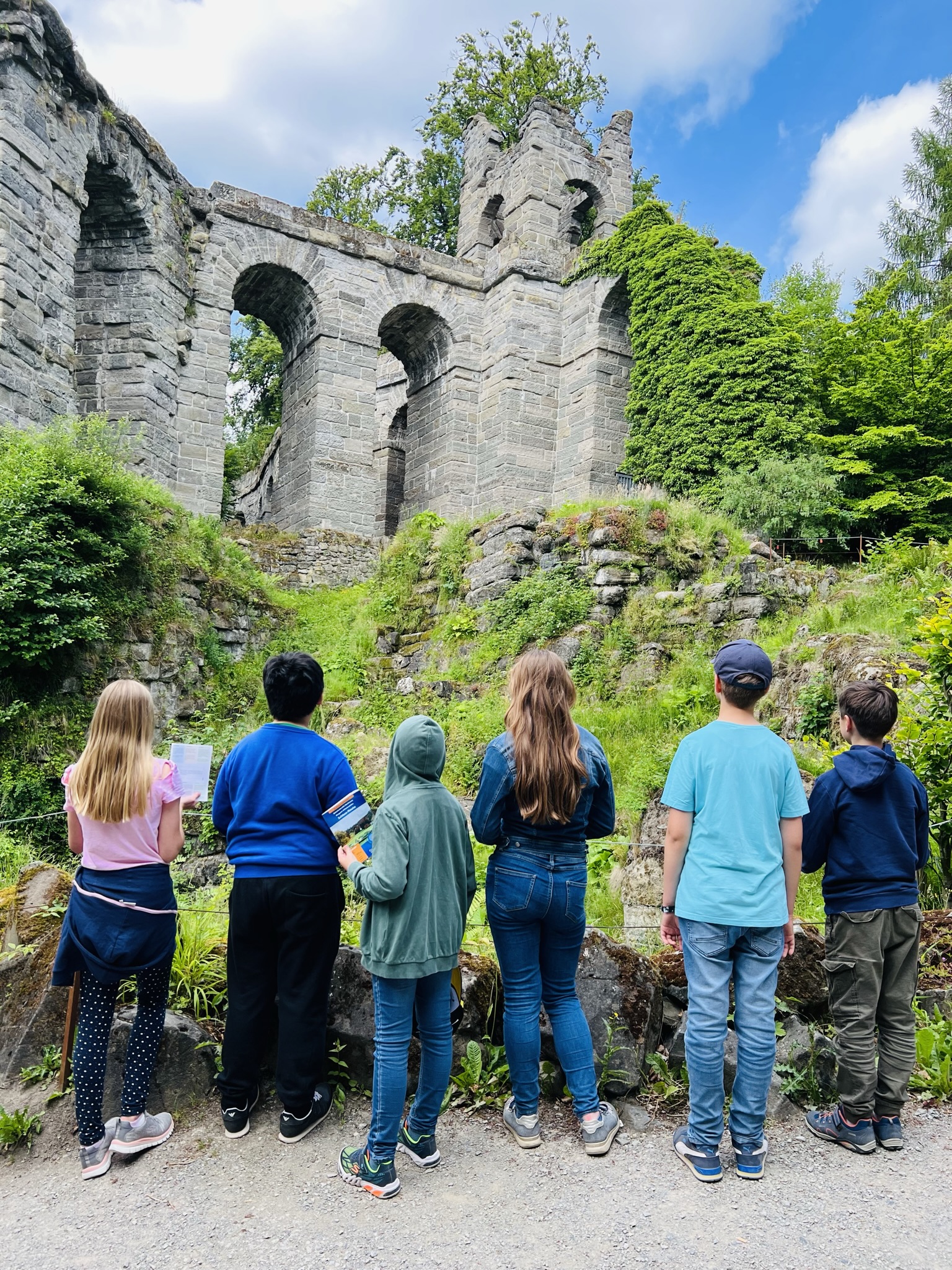 Kindergruppe bei den Stadtkrimis Kassel steht mit Rätselheften vor einer alten Steinruine im Bergpark, umgeben von Natur und sommerlicher Kulisse