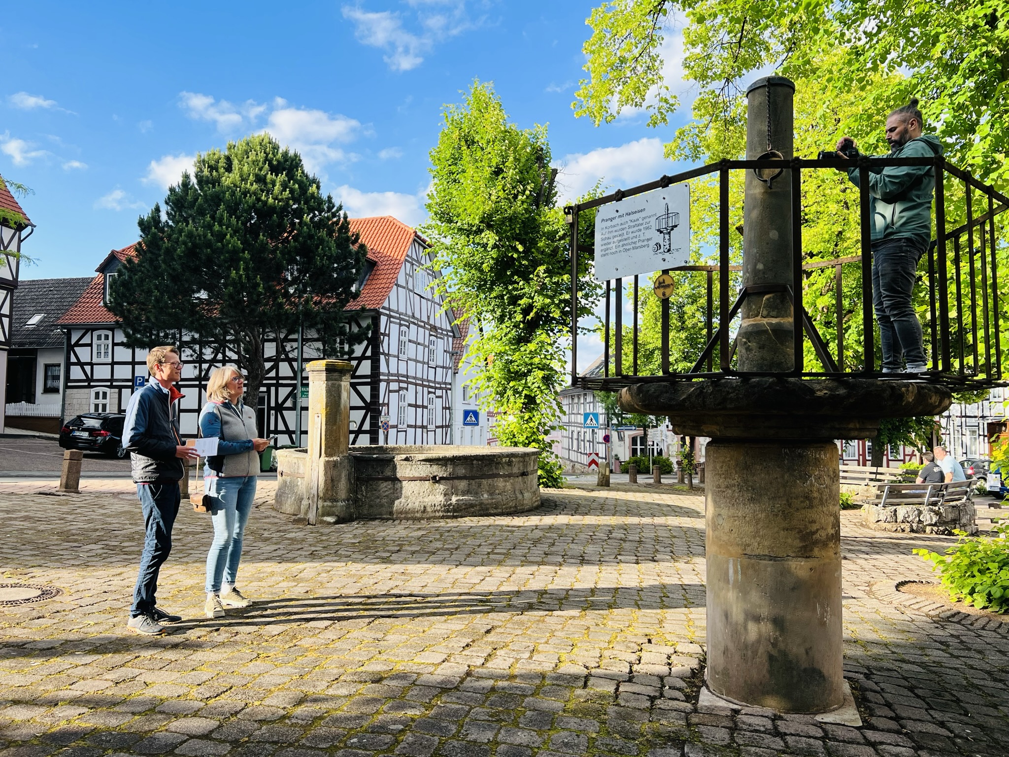 Zwei Stadtkrimi-Teilnehmende stehen an einem historischen Brunnenplatz mi Fachwerkhäusern im Hintergrund, bei sonnigem Wetter in Kassel auf Spurensuche