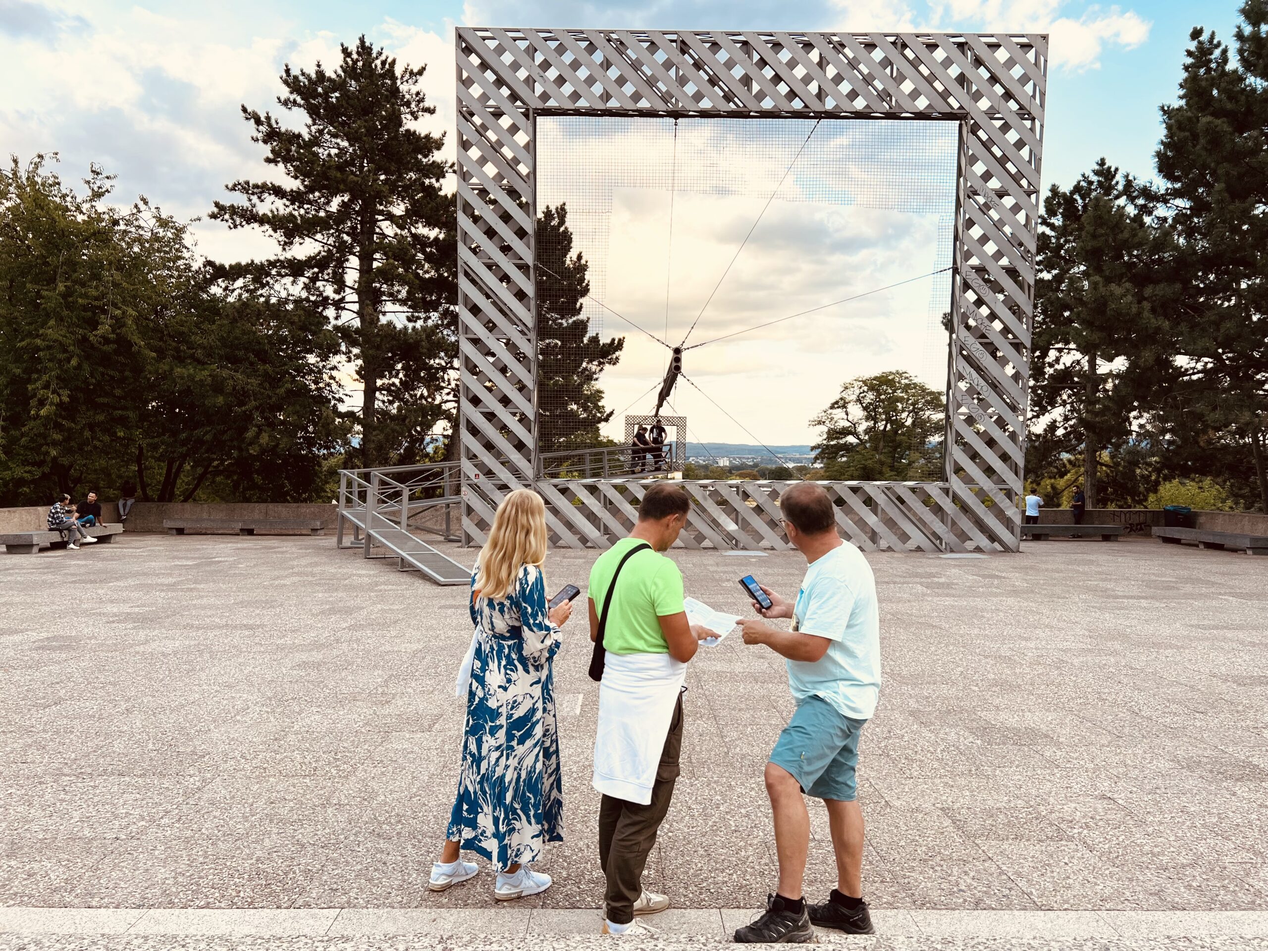 Drei Teilnehmer der Stadtkrimis Kassel stehen vor dem großen Himmelsscheibenrahmen an der Grimmwelt Kassel, mit Roadbook in der Hand und Blick auf das Panorama