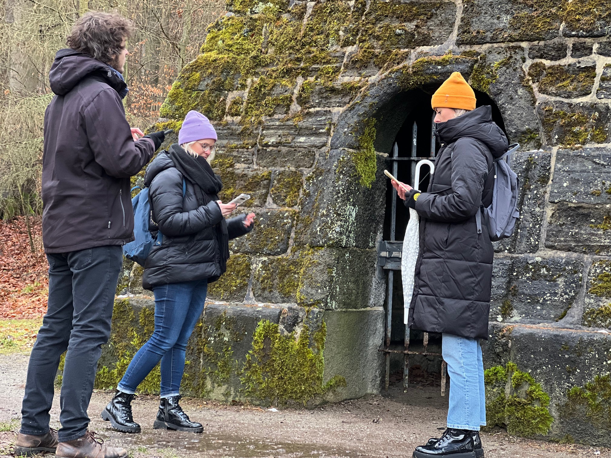 Drei Stadtkrimi-Teilnehmende stehen mit Smartphones vor einem moosbewachsenen alten Steingebäude mit Gittertor – rätselhafte Stimmung im Kasseler Bergpark