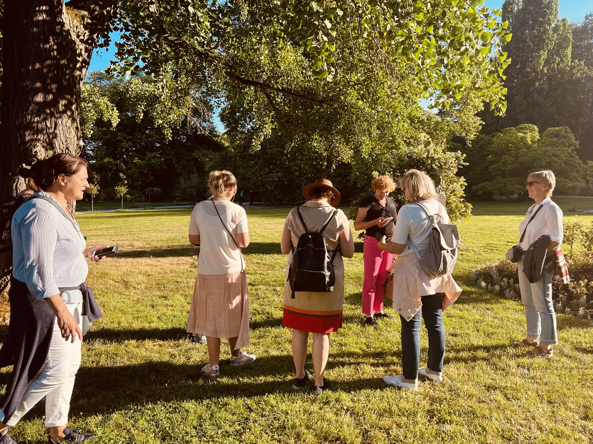 Gruppe von Stadtkrimi-Teilnehmenden im Park bei sonnigem Wetter, diskutieren über Hinweise mit Smartphone und Roadbook in der Hand unter Bäumen