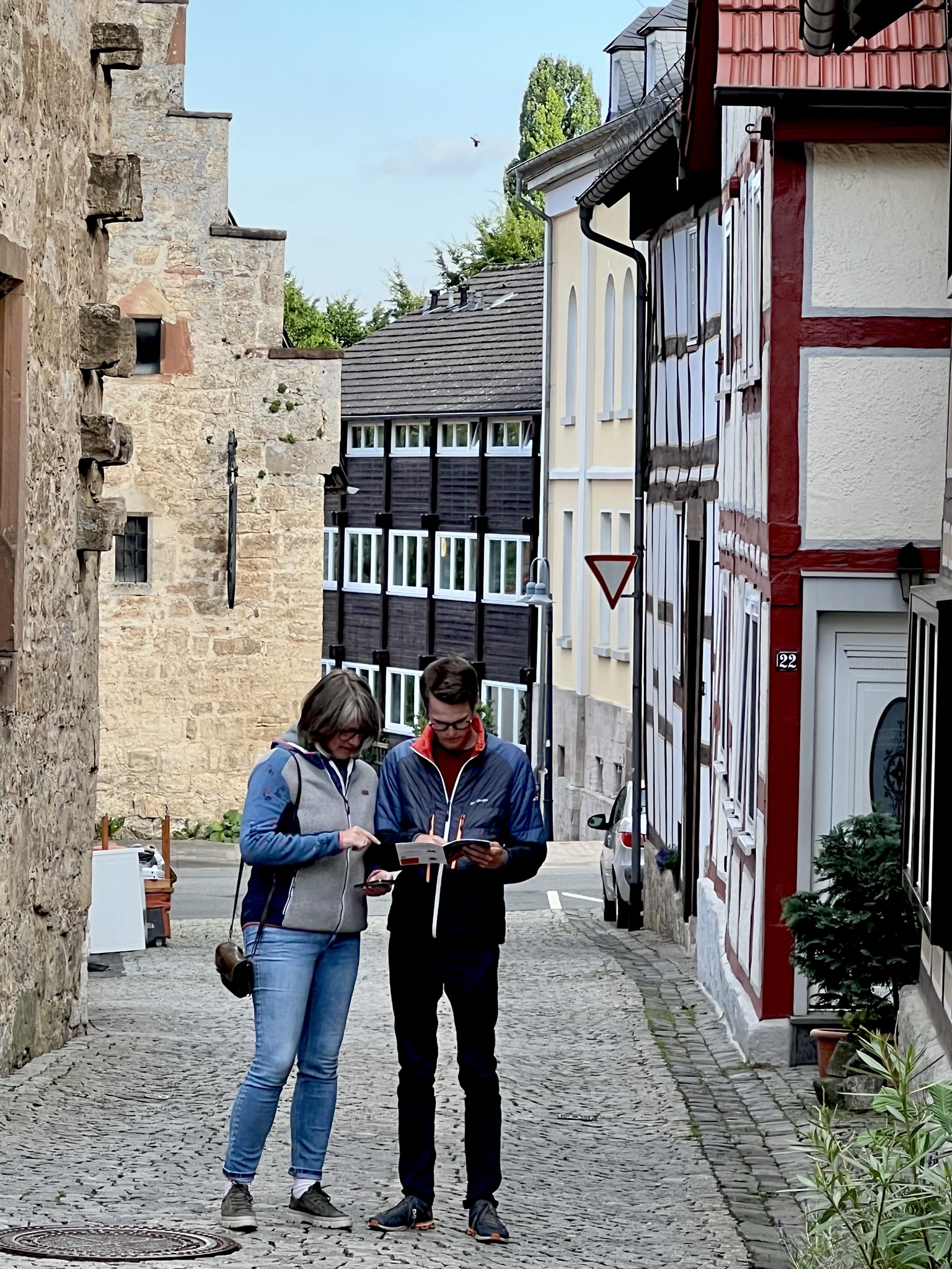 Zwei Teilnehmer der Stadtkrimis Kassel stehen mit einem Stadtplan oder Roadbook in einer historischen Gasse mit Fachwerkhäusern und Burgruine im Hintergrund