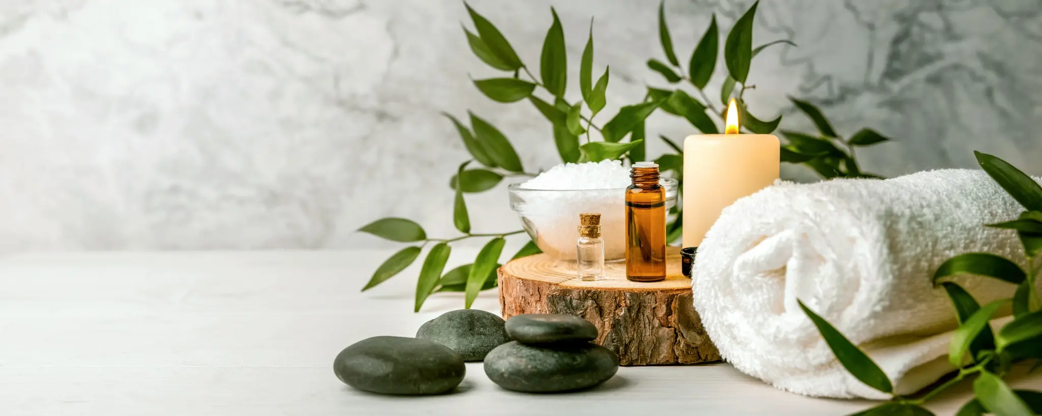 Wellness-Arrangement mit Massage-Steinen, ätherischen Ölen, Salz, Kerze und Blattdeko auf hellem Hintergrund