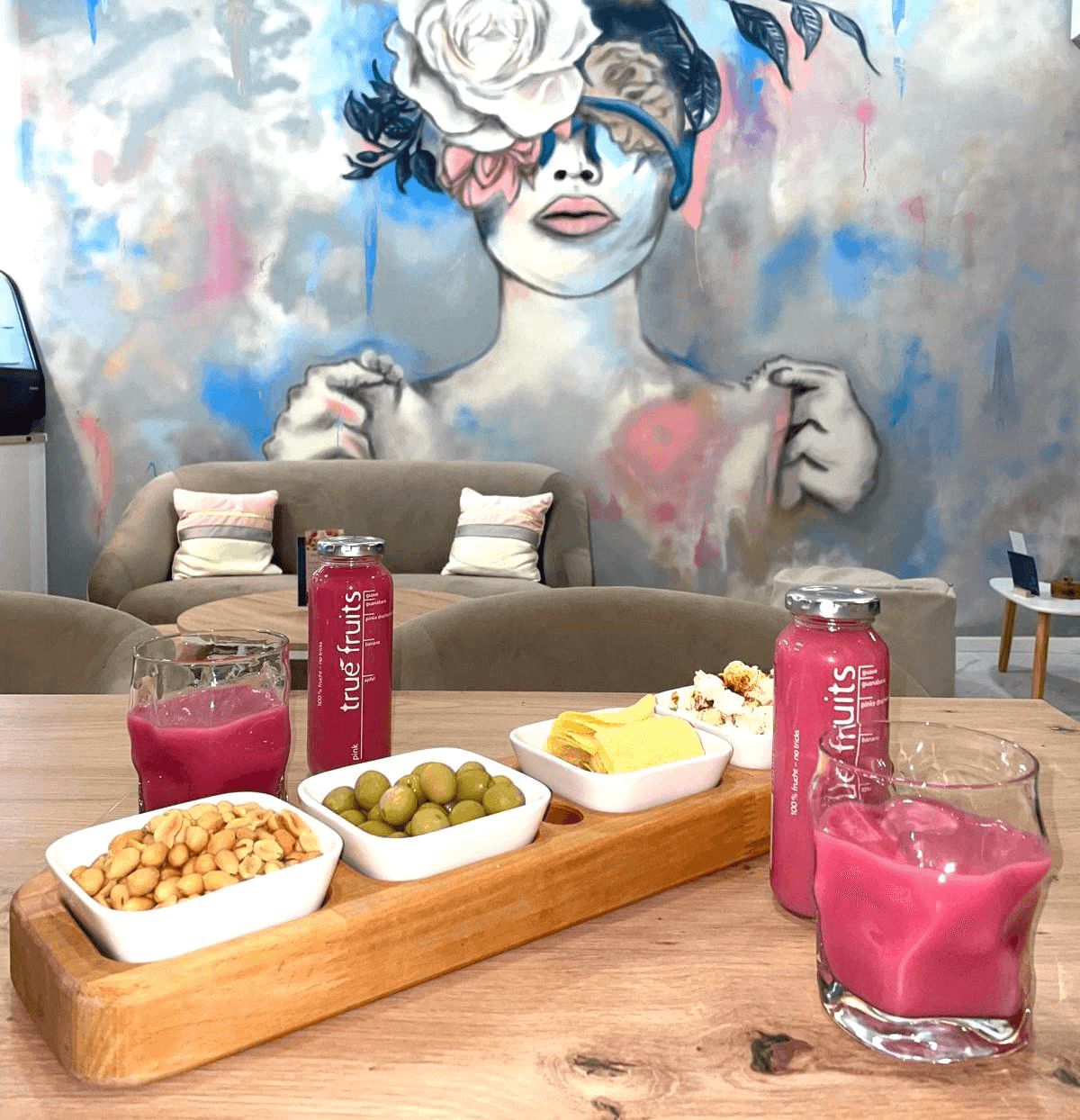 Serviertablett mit Oliven, Käse, Nüssen und pinkfarbenen Getränken auf einem Tisch vor einer Wand mit moderner Kunst einer stilisierten Frau mit Blumenkrone