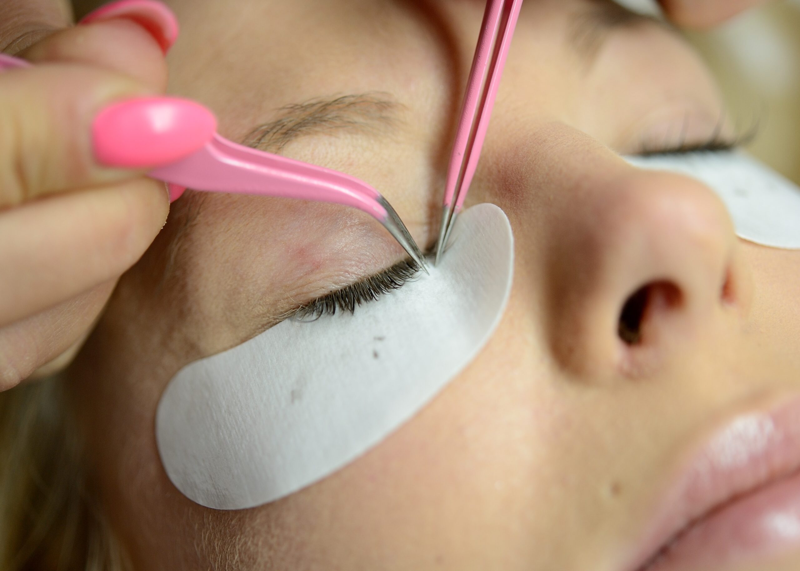 Nahaufnahme einer Wimpernverlängerung mit pinken Pinzetten, einzelne Extensions werden präzise aufgetragen, das untere Lid ist mit einem Schutzpad abgeklebt