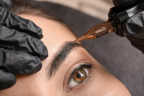 Nahaufnahme einer Augenbrauen-Pigmentierung mit Permanent-Make-up-Gerät durch eine Kosmetikerin mit schwarzen Handschuhen