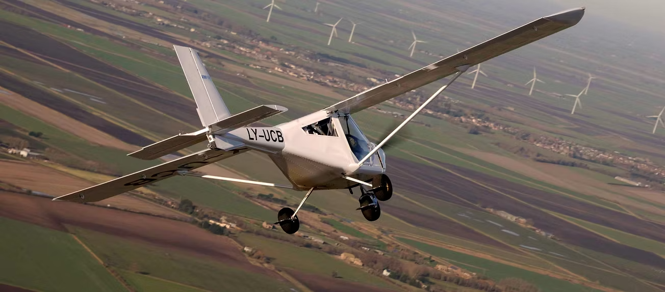 Weißes Flugzeug mit dem Kennzeichen LX-AJOB im Flug über Felder mit Windrädern im Hintergrund, aufgenommen in leichter Schräglage