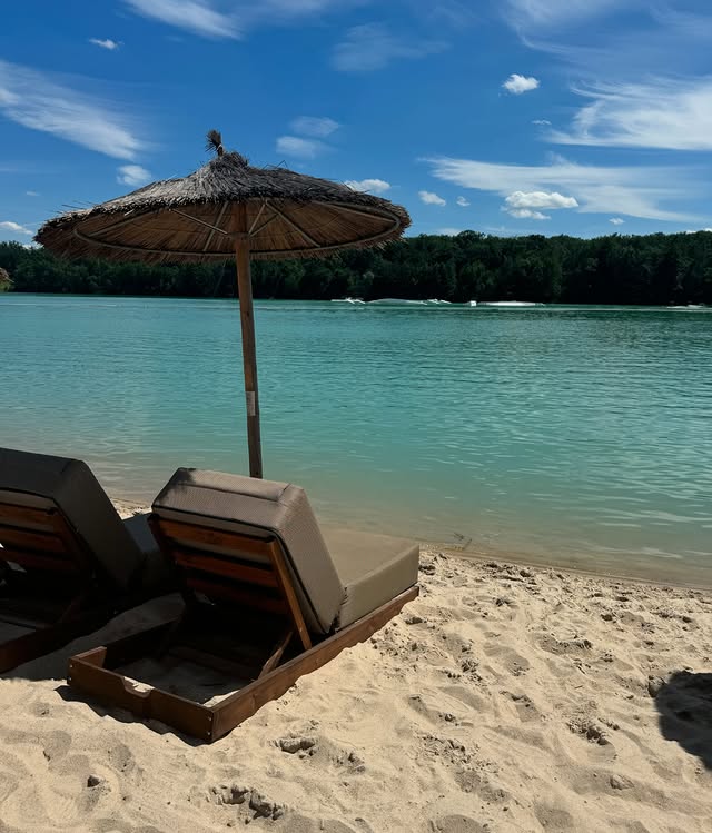 Zwei Liegestühle mit Strohdach direkt am Sandstrand des Chamäleon am See in Raunheim, mit Blick auf türkisfarbenes Wasser und bewaldetes Ufer