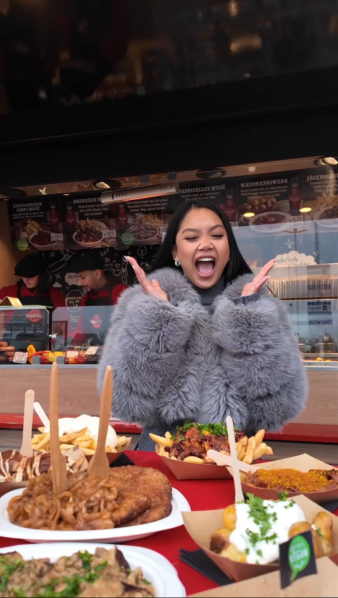 Frau in grauem Kunstpelz vor Foodtruck von Extrawurst mit verschiedenen deftigen Gerichten wie Currywurst, Pommes und veganem Menü