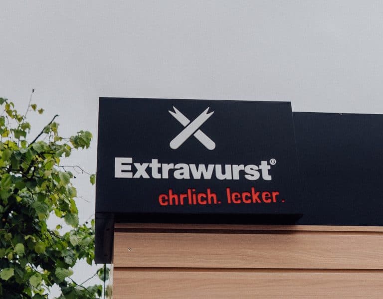 Logo von Extrawurst auf einem Filialenschild mit dem Claim „ehrlich. lecker.“ vor grauem Himmel