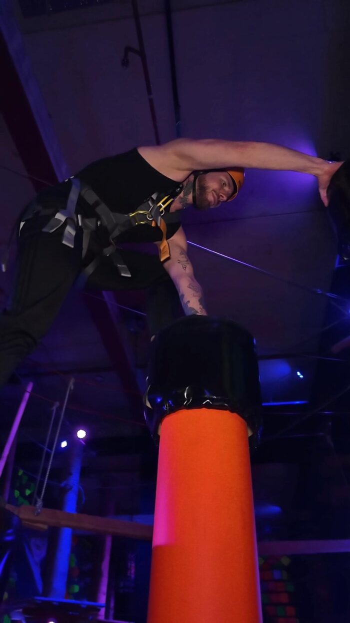 Mann mit Kletterausrüstung balanciert auf einem hohen Kletterblock, lila und blaue Lichtstimmung