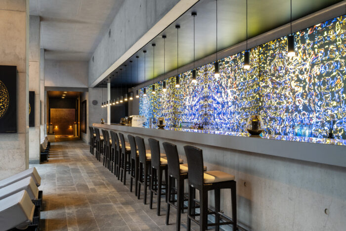 Designorientierter Barbereich mit hohen Hockern, schwebenden Pendelleuchten und einer hinterleuchteten Wand aus farbigem, abstrakt schimmerndem Mosaik in Blau-, Gelb- und Weißtönen.