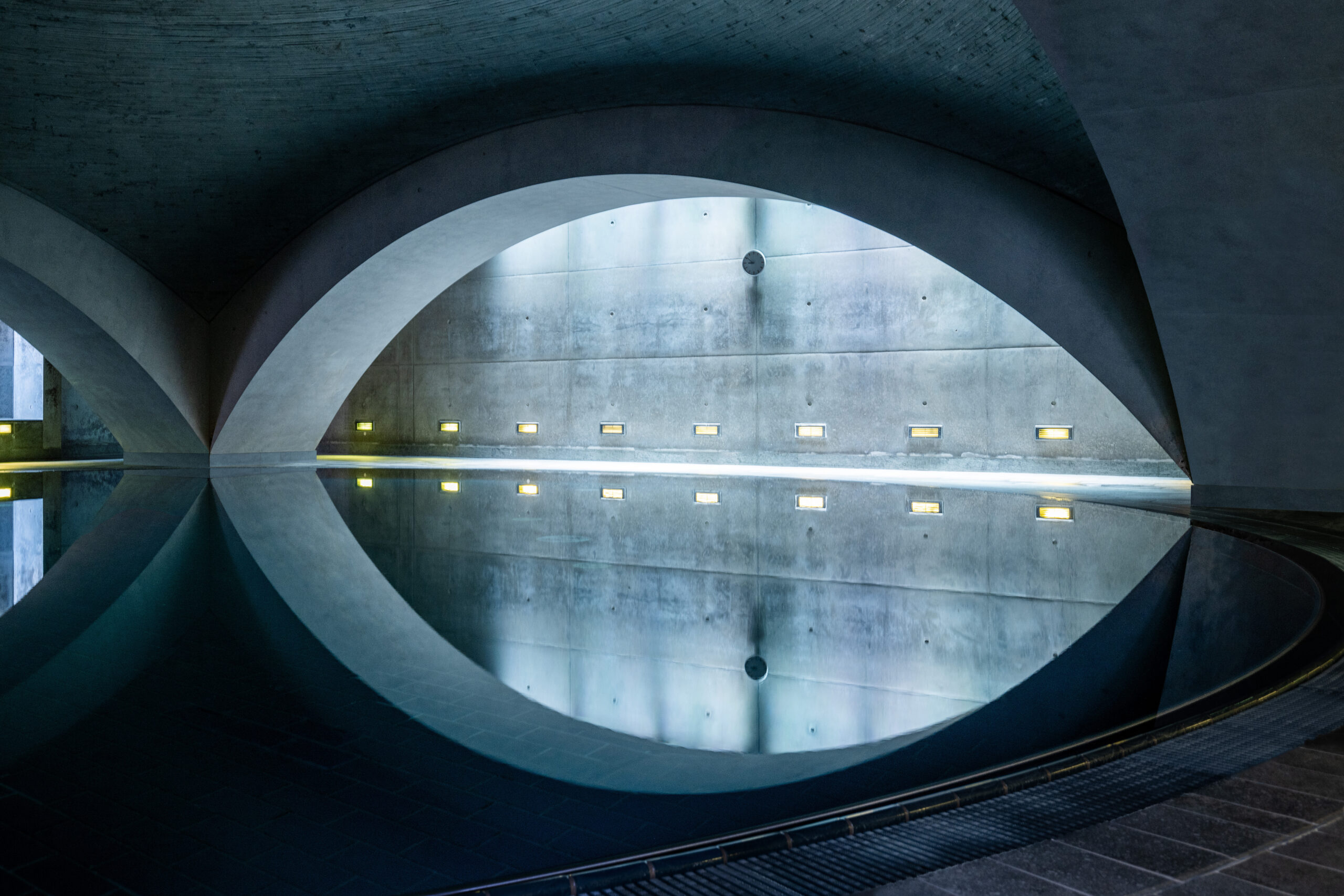 Detailaufnahme eines gewölbten, architektonisch eindrucksvollen Innenraums mit spiegelglattem Wasserbecken und sanfter Beleuchtung an einer Sichtbetonwand.