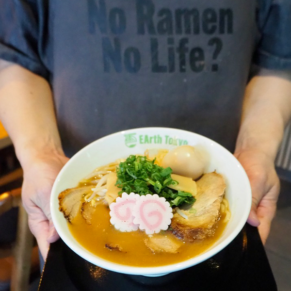 Mann in „No Ramen No Life“-Shirt hält eine Schale Ramen mit Fleisch, Narutomaki und frischem Gemüse
